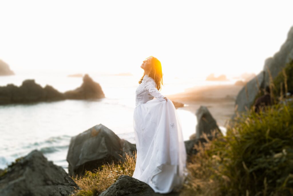 women in white dress with her arms open with the sun shining on her face. Standing by the ocean. 